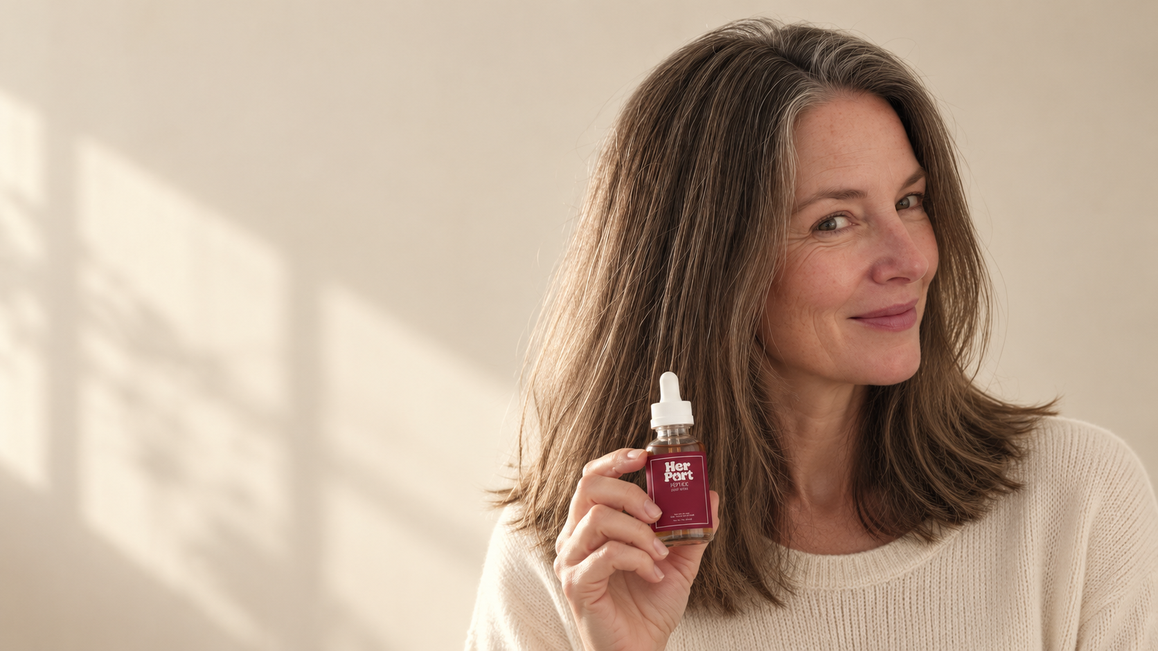 Woman in her fifties with full brown-and-silver hair holding her Her Part serum
